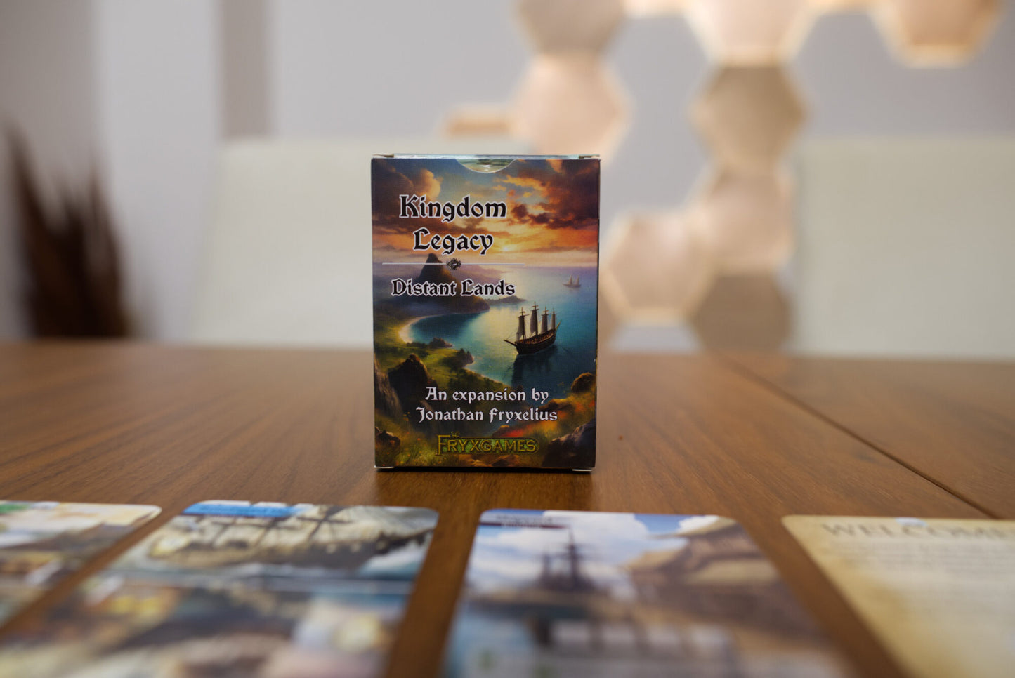 Kingdom Legacy: Distant Lands board game box standing on a wooden tabletop, with several cards blurred in the foreground and a softly lit indoor background.
