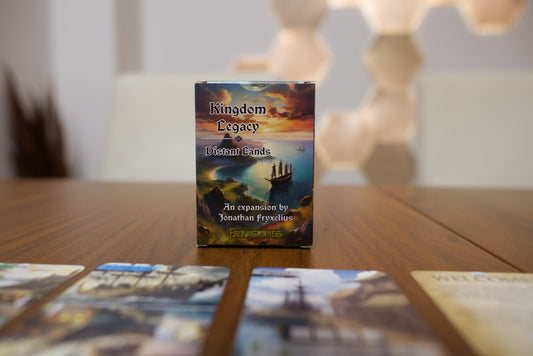 Kingdom Legacy: Distant Lands board game box standing on a wooden tabletop, with several cards blurred in the foreground and a softly lit indoor background.