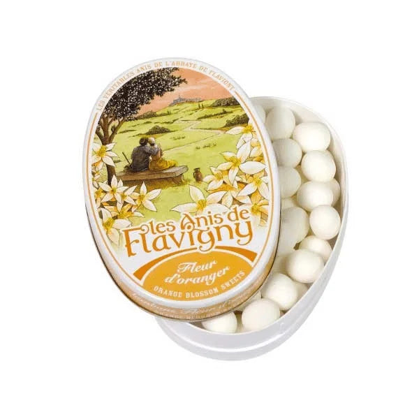 A partially open 50g tin of Twice as Sweet Anis De Flavigny Orange Blossom candies reveals small white French sweets. The lid shows a countryside picnic with orange blossoms. Made in France.