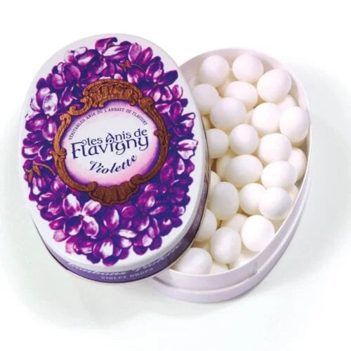 A partially open 50g oval tin of Twice as Sweet Anis De Flavigny Violette features purple violets on the lid and reveals small round violet candies inside. Product of France.