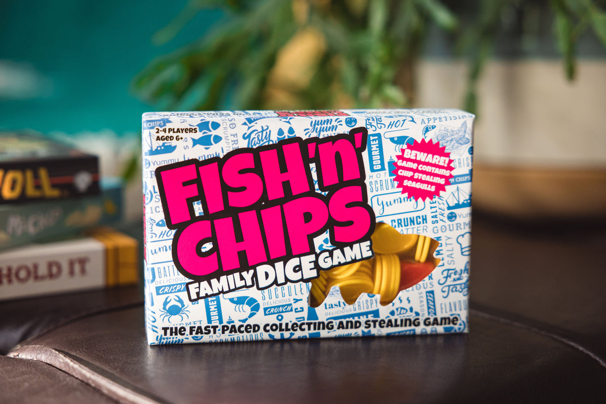 The product box for 'Fish 'n Chips Family Dice Game' is shown on a dark wooden table with a blurred background of books and greenery.