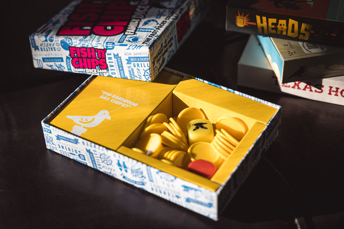 An open box of for the family dice game "Fish & Chips" on a dark wooden surface, with other branded game boxes in the background.