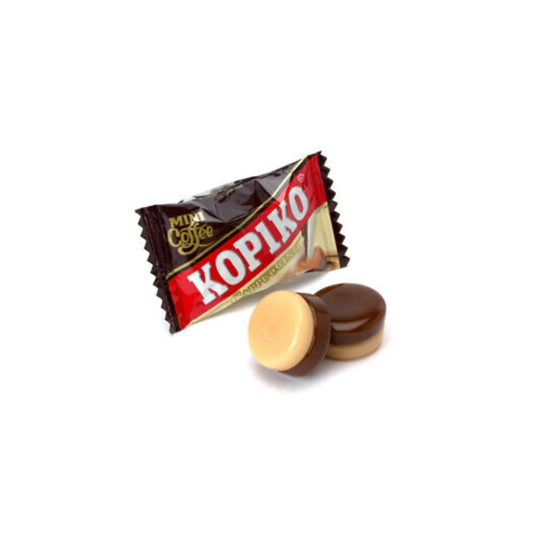 A Twice as Sweet Kopiko Cappuccino Candy 175g (Indonesia), in its brown and cream wrapper, sits beside two unwrapped, two-tone candies—half cream, half brown—on a white background, showcasing this popular coffee treat.