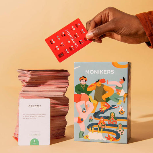 Hand holding a red card above a box of 'Monikers' game on a beige background