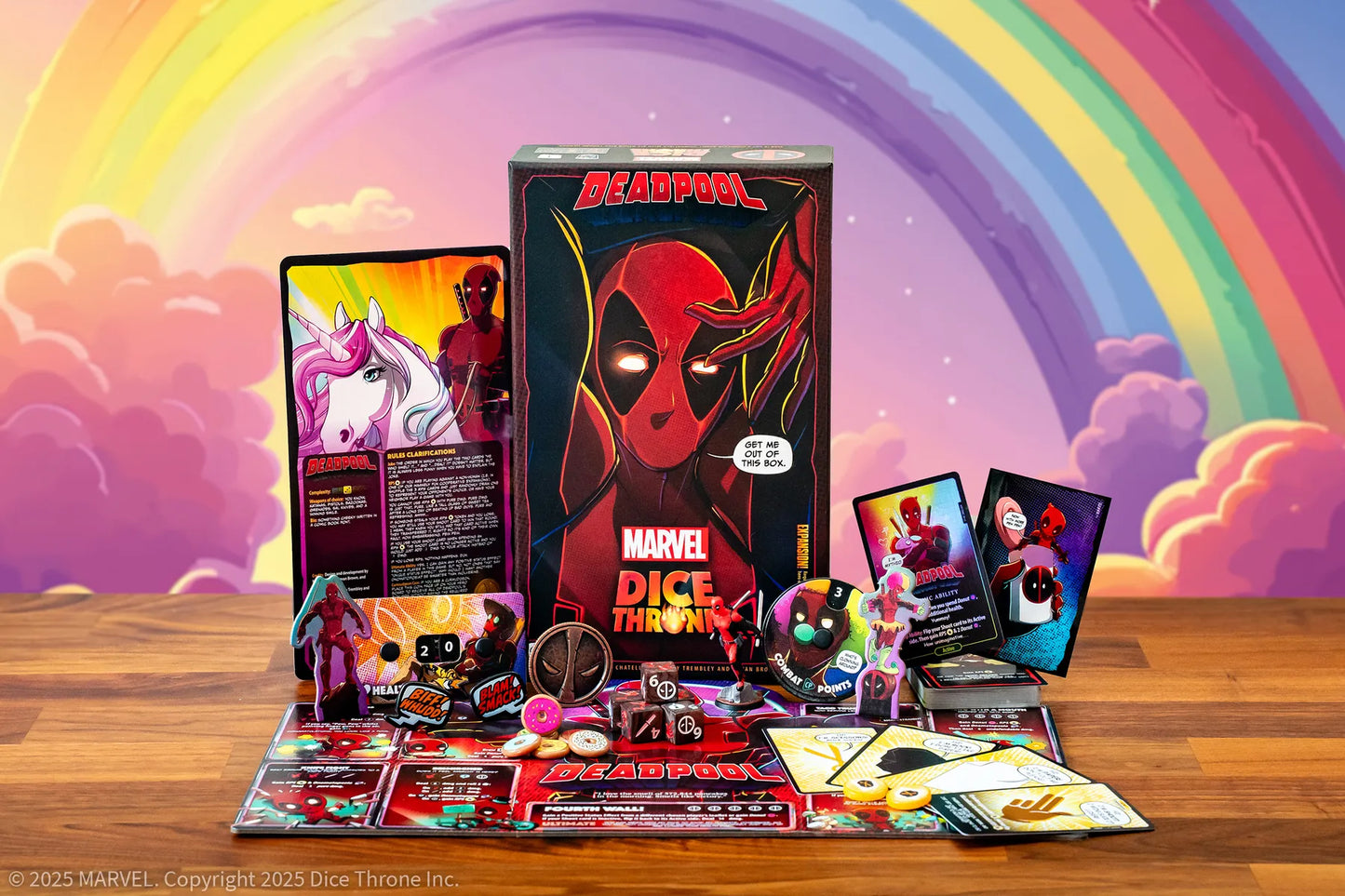 Deadpool Dice Throw board game set displayed on a wooden table with its box, cards, dice, and accessories arranged in front. The background features a colorful pastel rainbow and clouds.