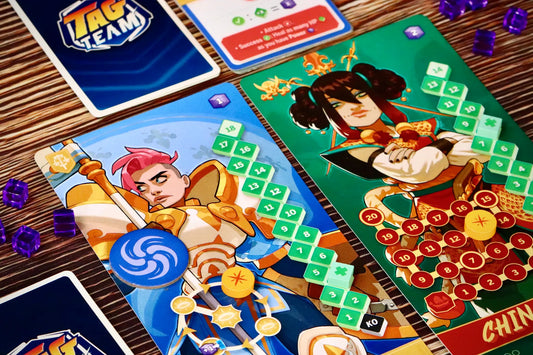 Close-up of a board game setup on a wooden table, showing illustrated playmat cards with colorful fantasy characters, game tokens, purple dice, and part of a blue card labeled TAG TEAM.
